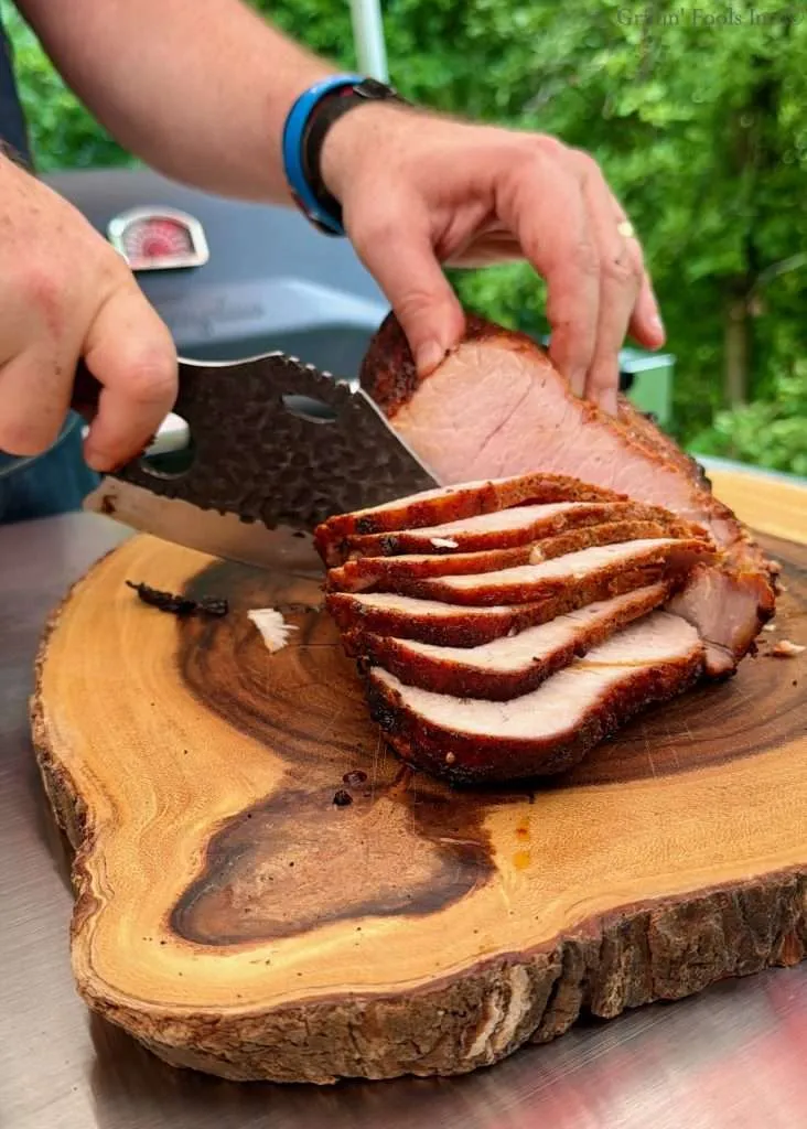 Close-up of a wicked looking knife slicing rotisserie pork loin on a round wooden cutting board outdoors.