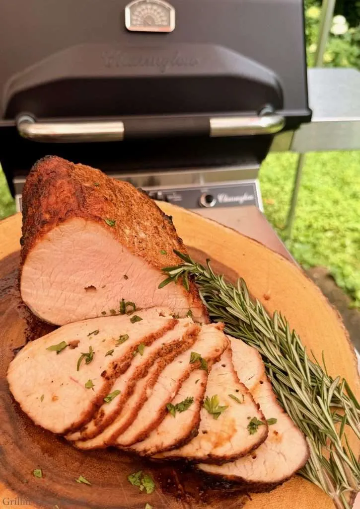 Sliced roasted pork on a cutting board with fresh rosemary sprigs, grill in the background outdoors.