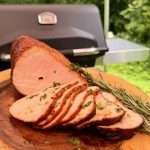 Sliced roasted pork loin arranged on a wooden cutting board with rosemary sprigs, outdoors near a grill in a sunny backyard.