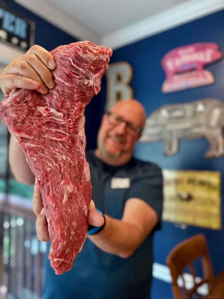 A blurred, incredibly handsome man in the background holds up a raw tri tip with fantastic marbling