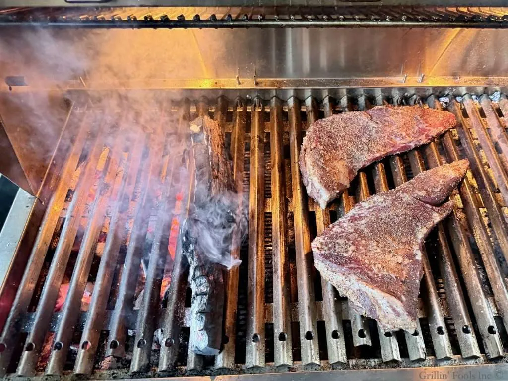 Two seasoned tri tip roasts, next to two chunks of wood, smoking over hot coals on a BBQ Grill