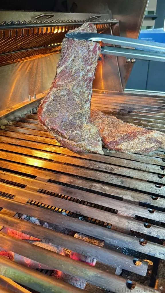 A partially cooked beef tri tip, being held up by tongs as it makes its way to be dropped over the hot coals