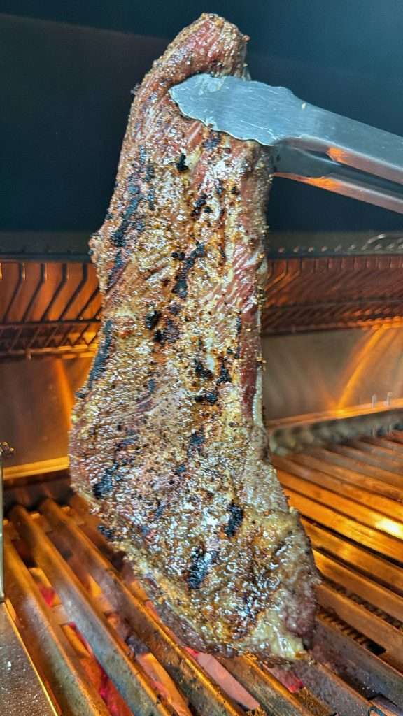 Tongs holding a charred tri tip up over the hot coals on a charcoal grill