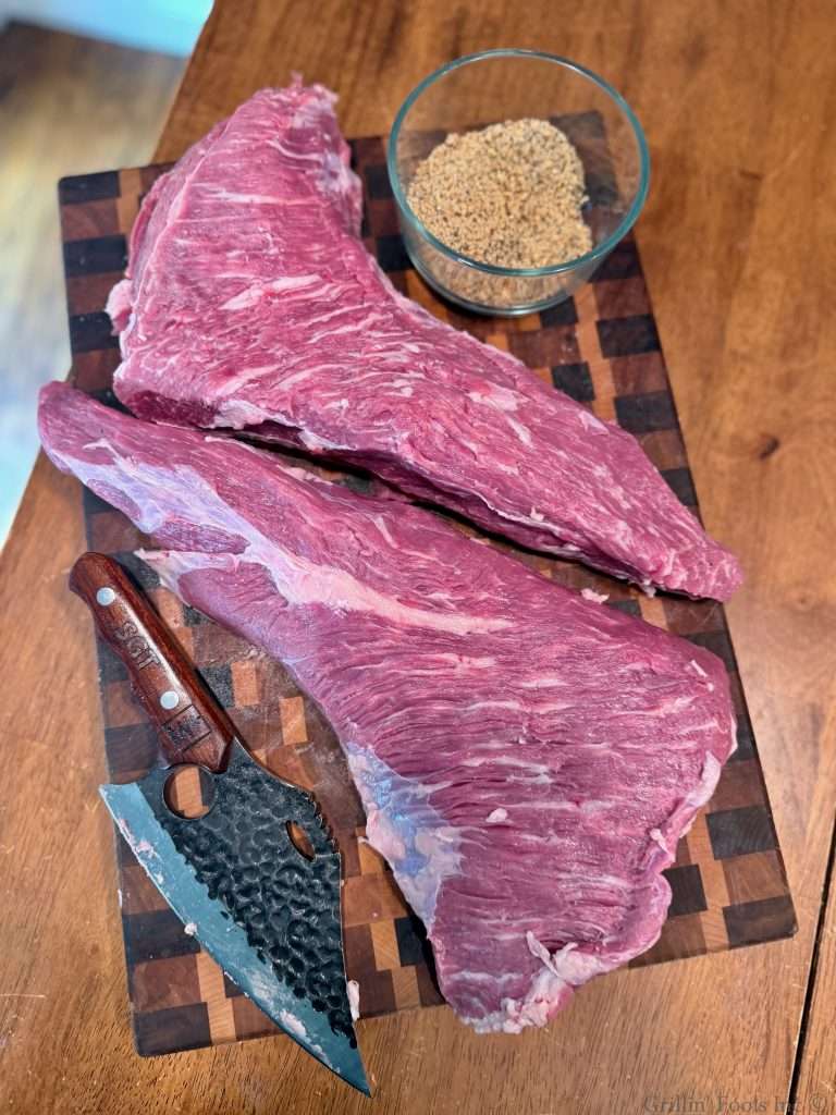 Two tri tip roasts on a cutting board with a bowl of seasoning and a wicked looking knife, taken from above.