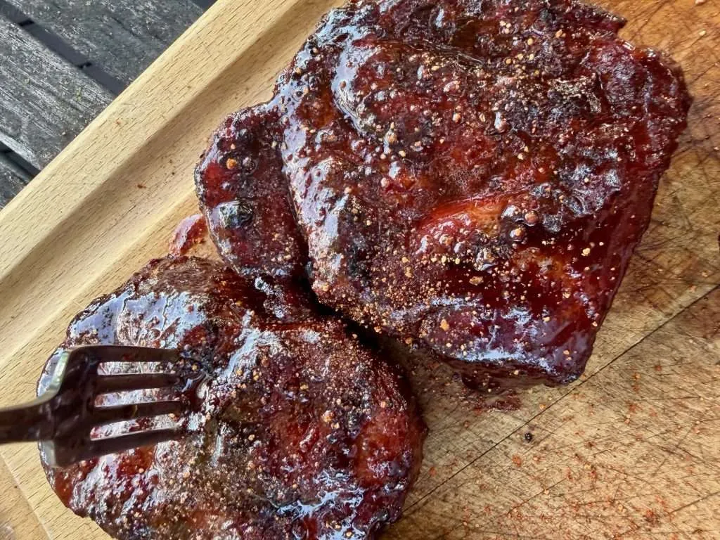 A couple sauced and seasoned pork collars from above on a wooden cutting board with a fork stuck into one.