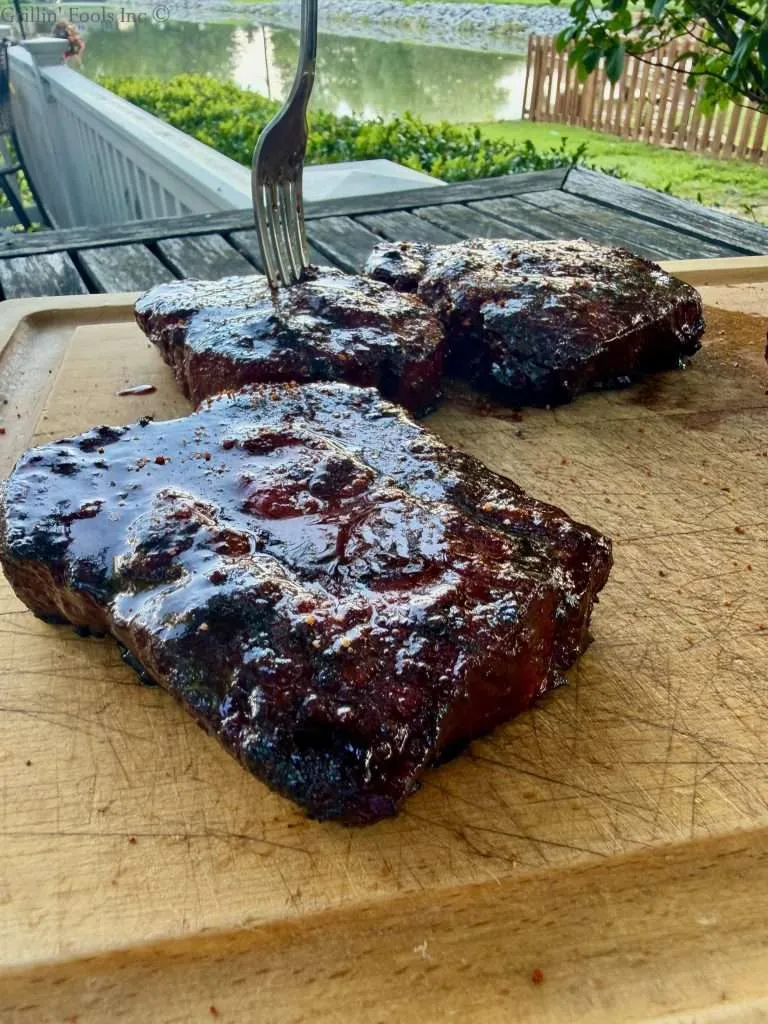 Saucy pork collar, dusted with BBQ rub on a wooden cutting board with a lake in the background