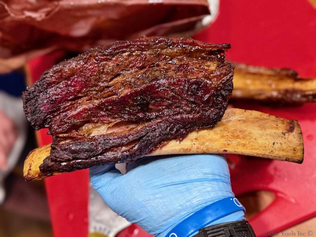 English Cut Smoked Beef Ribs being held in a light blue gloved hand over a red cutting board English Cut Smoked Beef Ribs being held in a light blue gloved hand over a red cutting board