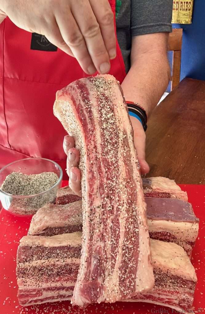 A man holds a well marbled, raw beef rib and applies a beef seasoning to one side