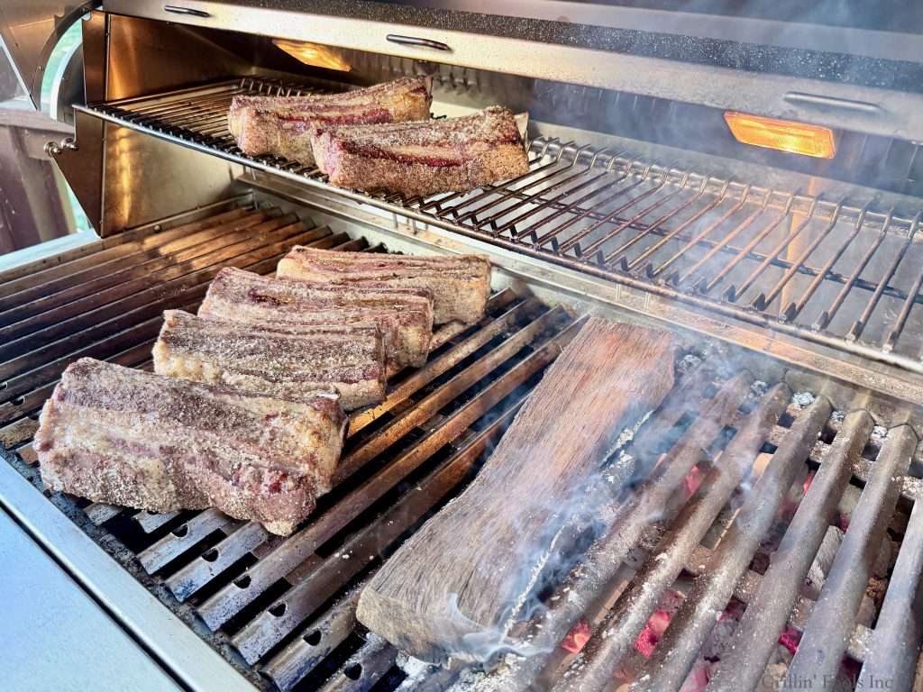 Seasoned beef ribs on a charcoal grill, next to a smoking oak log over hot coals