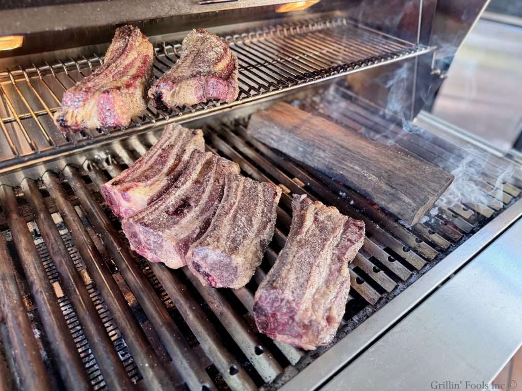 Seasoned beef ribs on a charcoal grill, next to a smoking oak log over hot coals