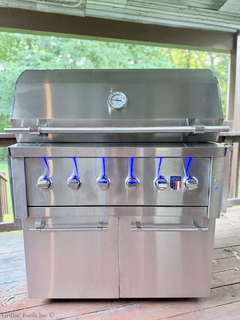 The American Made Grills Hybrid Ovation Grill on a deck with green foliage in the background