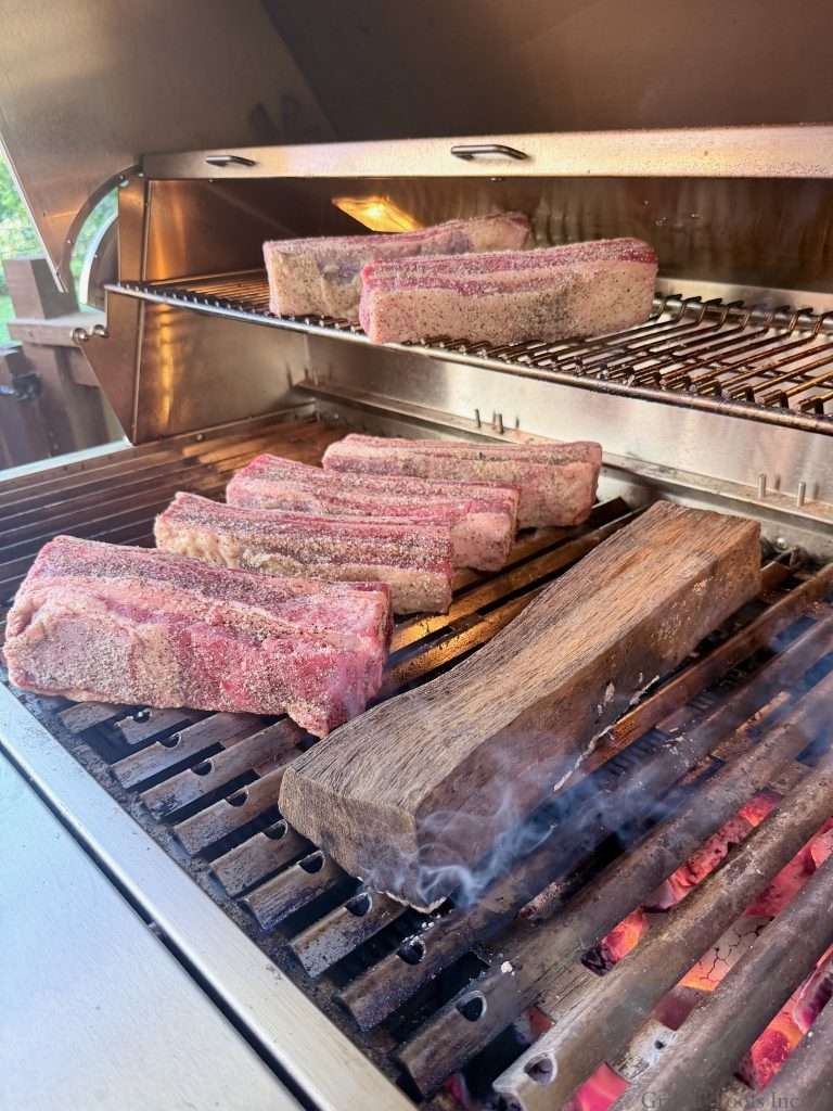 Seasoned beef ribs on a charcoal grill, next to a smoking oak log over hot coals