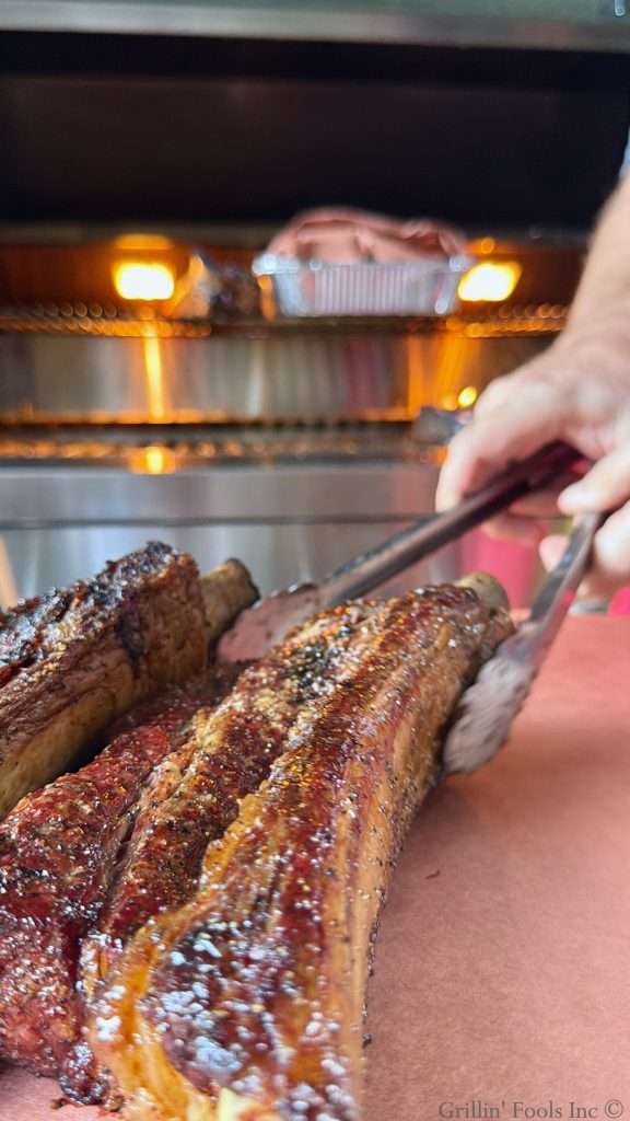 A man moves beef ribs with a pair of tongs from the grill to a sheet of peach butcher paper