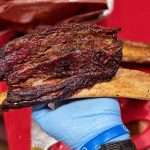 A smoked beef rib with glorious bark on a light blue gloved hand over a red cutting board