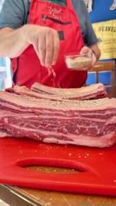 A man in a red apron seasons English cut beef ribs with SPG rub