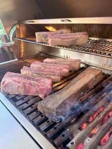 Seasoned beef ribs on a charcoal grill, next to a smoking oak log over hot coals