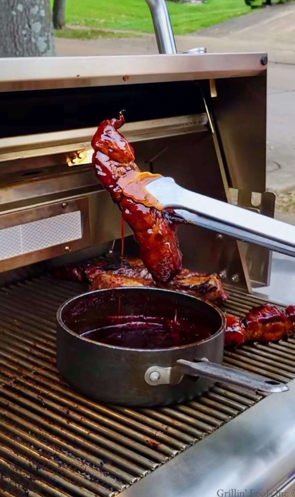 A country style rib, held up by a set of tongs, with BBQ sauce running off it into a pan of BBQ sauce