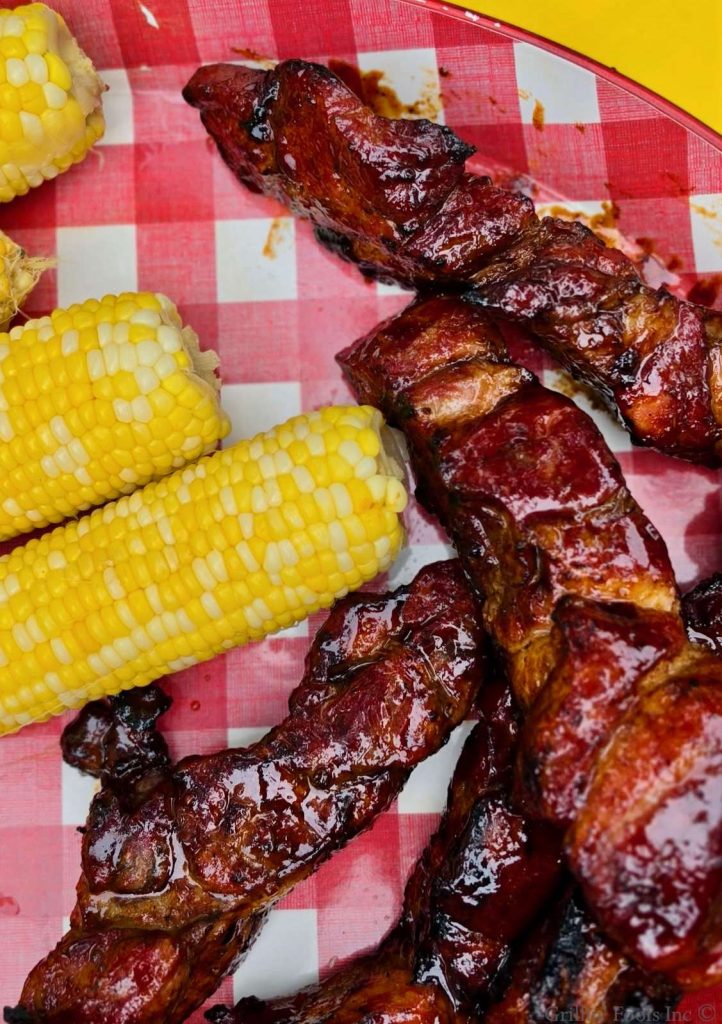 Saucy country style ribs on a red and white checked platter with fresh yellow corn