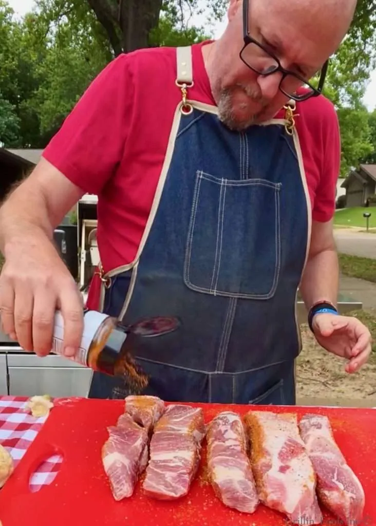 A devastatingly handsome man in a red shirt and denim apron seasons country style ribs