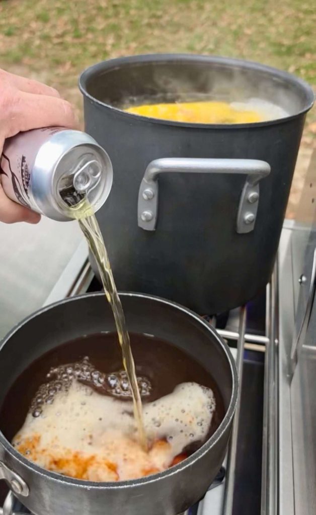 Beer pours into a pan of BBQ sauce in front of a pot holding boiling water and corn.