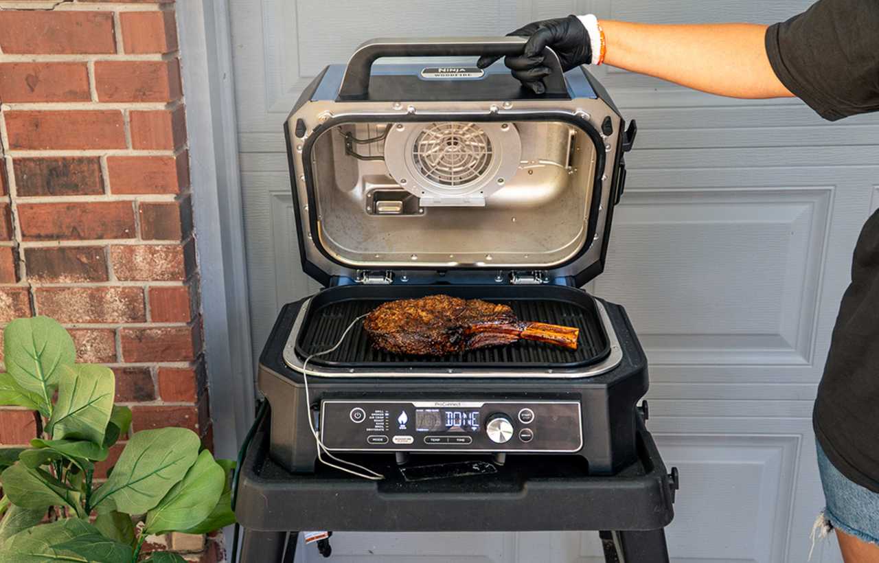 A cooked tomahawk steak rests on the grill grate of a Ninja Woodfire Pro XL, with the lid open to reveal the interior heating fan. A hand wearing a black glove holds the lid open, while the grill sits on a black cart in front of a garage door and red brick wall.