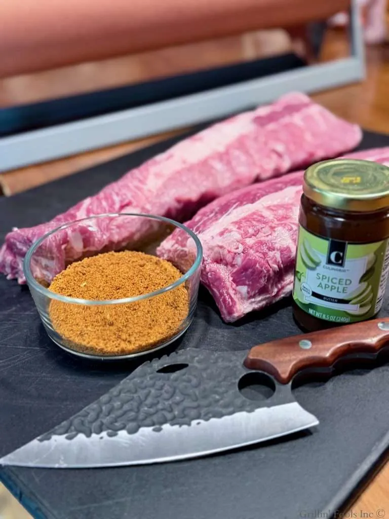 Two raw slabs of baby back ribs, a bowl of seasoning, jar of apple butter, and a knife on a black cutting board, with a roll of pink butcher paper in the background