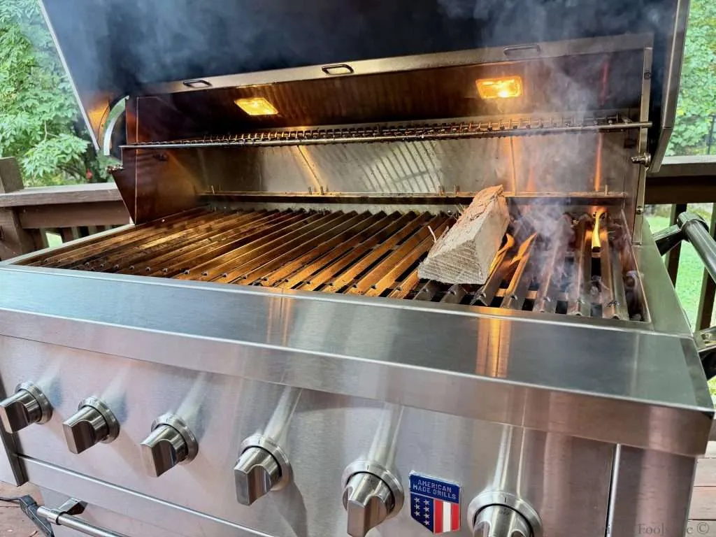 An American Made Grills, Hybrid Ovation Grill with hot coals on the right side and a hickory wood split just off the edge of those hot coals