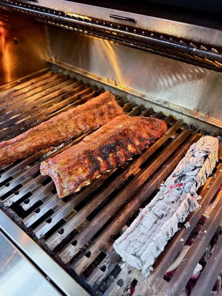 A slab of baby back ribs with a little char on one side next to hickory split that is reduced to ash