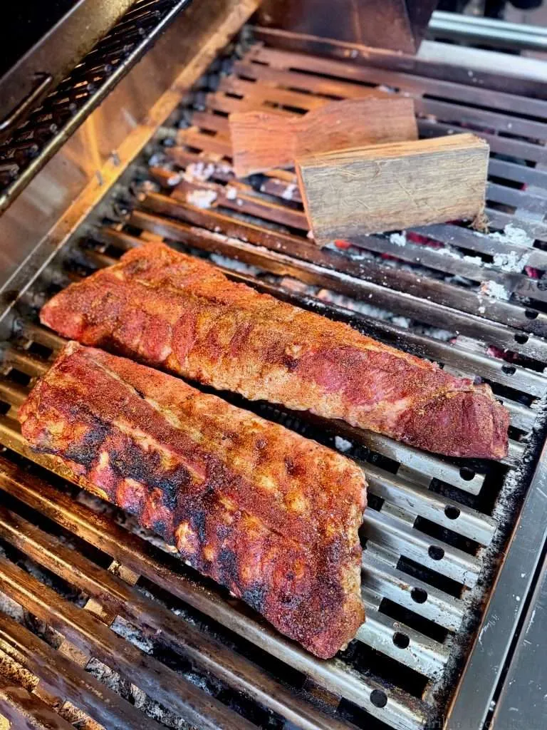 Two slabs of baby back ribs, repositioned next to a couple small chunks of fresh smoke wood