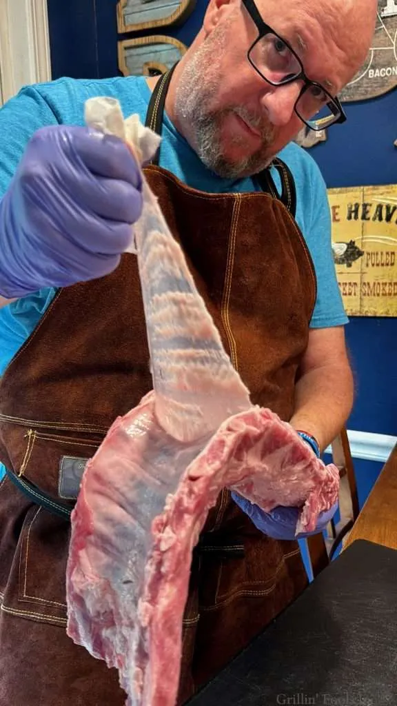 A devastatingly handsome bearded man, in light blue nitrile gloves pull the membrane off the back of a slab of baby back ribs