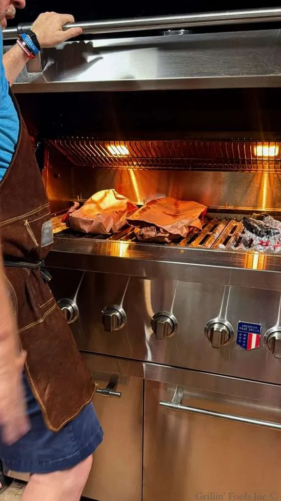 A man in a brown suede apron stands near a BBQ grill with two slabs of ribs wrapped in butcher paper