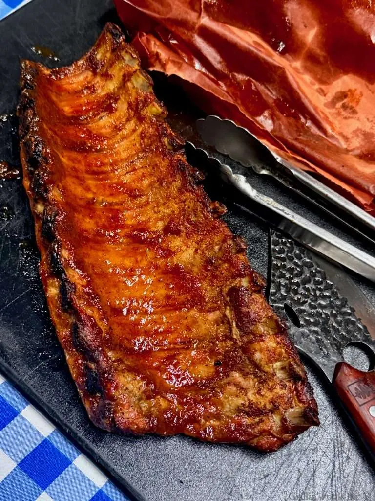 A slab of smoke baby back ribs with apple butter next to a wicked knife, tongs, wet butcher paper all on a black cutting board