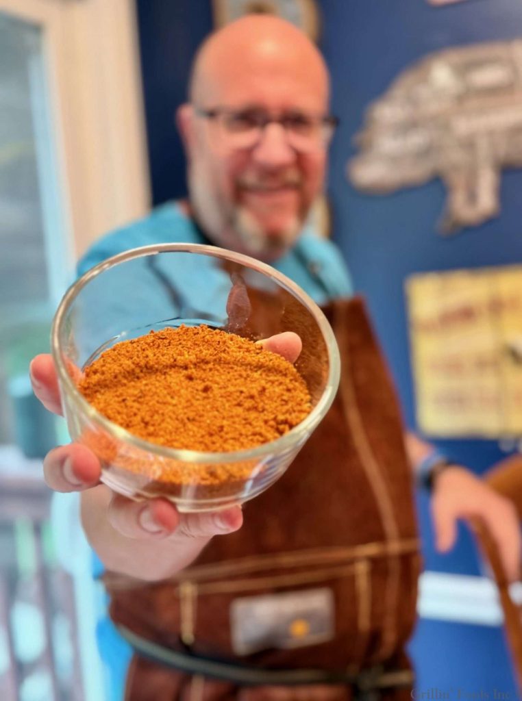 A gorgeous man in a brown suede apron holds a bowl of BBQ rub